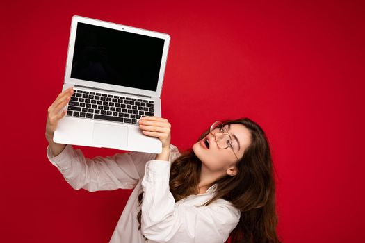 Photo shot of amazing funny happy beautiful smiling dark hair young woman holding computer laptop with empty monitor screen with mock up and copy space wearing white shirt looking up with open mouth isolated over red wall background.