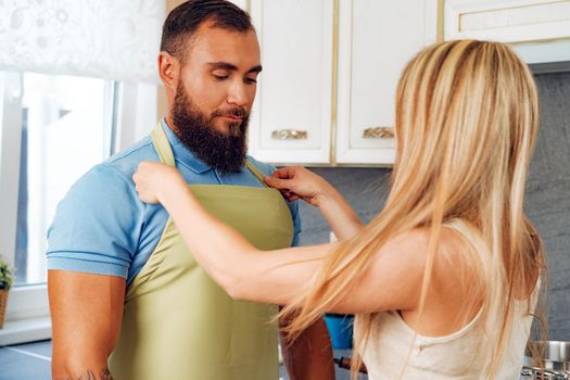 Blonde woman putting apron on her husband in their kitchen close up