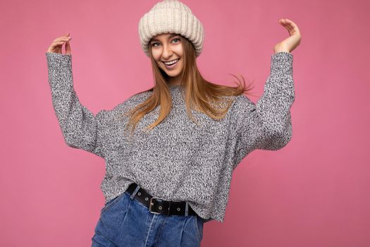 Photo of beautiful positive joyful young dark blonde woman standing isolated over pink background wall wearing grey sweater and winter beige hat looking at camera dancing and having fun.