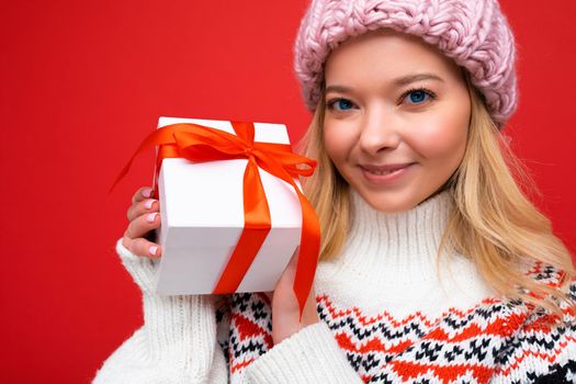 Beautiful happy young blonde woman isolated over colourful background wall wearing stylish casual clothes holding gift box and looking at camera.