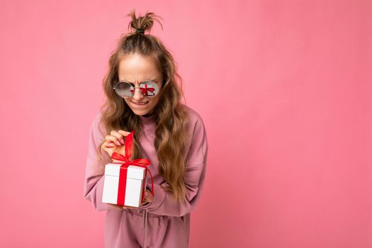 Shot of beautiful positive young blonde curly woman isolated over pink background wall wearing pink sport clothes and sunglasses holding gift box and unboxing surprise looking at present. Empty space