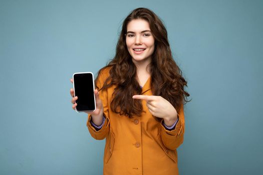 Photo of beautiful smiling young woman good looking wearing casual stylish outfit standing isolated on background with copy space holding smartphone showing phone in hand with empty screen display for mockup pointing at gadjet looking at camera.