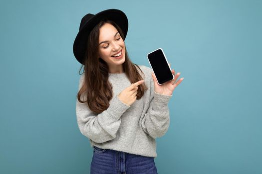 Closeup of charming young happy woman wearing black hat and grey sweater holding phone looking at mobile pointing finger at screen isolated on background.Mock up, cutout