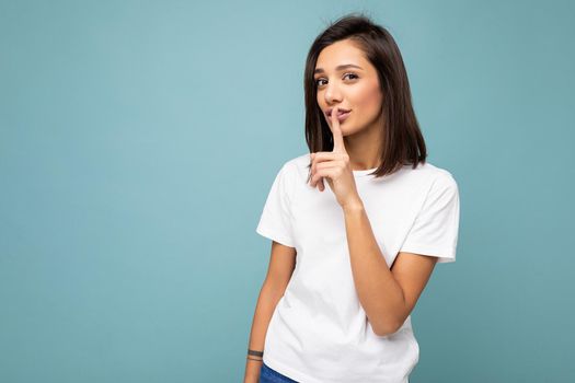 Portrait of young attractive brunette woman with sincere emotions wearing casual white t-shirt for mockup isolated on blue background with copy space and asking remain silent.