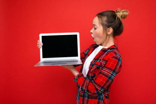 side profile photo of Beautiful shocked amazed young woman holding laptop wearing red shirt looking at monitor display isolated on red background. Cutout, free space