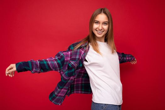 Photo of beautiful happy smiling young dark blonde woman isolated over red background wearing colourful stylish shirt and casual white t-shirt looking at camera and having fun.