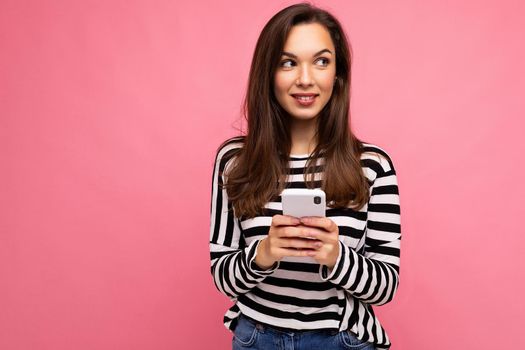 shot of pretty young smiling brunette woman using mobile phone communicating via texting sms wearing sweater isolated on wall background looking to the side. Copy space