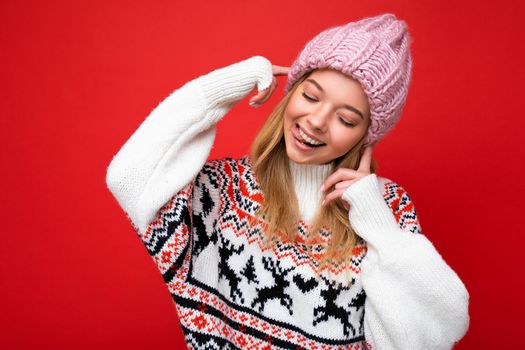 Photo of beautiful happy funny joyful young blonde woman isolated over red background wall wearing winter sweater and pink hat looking down and having fun.