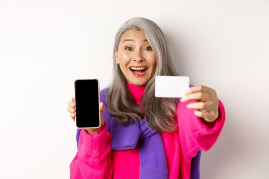 Online shopping. Close-up of stylish korean senior woman showing empty mobile screen and plastic credit card, smiling happy, standing over white background.