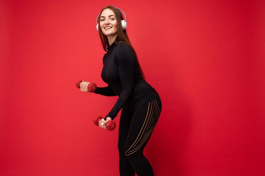 Side-profile photo of attractive positive smiling young brunette woman wearing black sport clothes isolated on red background wall doing fitness using dumbbells wearing white bluetooth earphones listening to good music looking at camera.