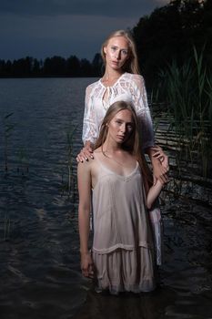 two young twin sisters with long blond hair posing in light dresses in water of lake at summer night, looking at camera. stylish fashion photoshoot with flashlight. outdoors evening photosession