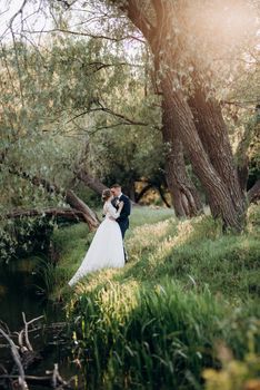 the groom and the bride are walking in the forest near a narrow river on a bright day