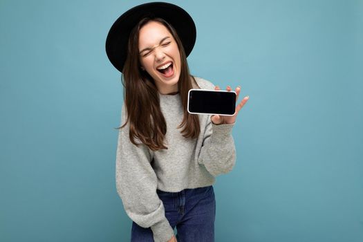 Portrait photo shot of beautiful young woman wearing black hat and grey sweater holding phone showing smartphone isolated on background with open mouth having joy.Mock up, cutout, copy space