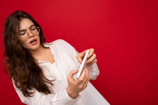 photo of Pretty smiling positive young brunette woman wearing white shirt and optical glasses isolated over red background holding in hand and using mobile phone playing online games looking at gadjet display.