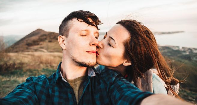 Young loving couple taking self-portrait on peak of mountain. Woman kissing a man with closed eyes