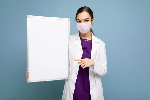Photo of young attractive nurse in protective face mask and white medical coat holding an empty magnetic board isolated on blue background.