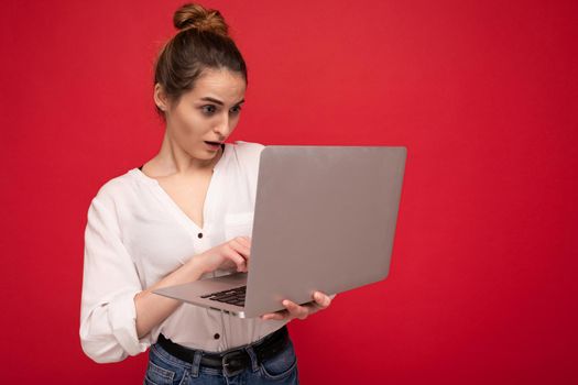 Side profile of Beautiful amazed brunet young woman holding netbook computer looking down with open mouth wearing white shirt typing on keyboard isolated on red background.