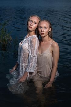 two young twin sisters with long blond hair posing in light dresses in water of lake at summer night, looking at camera. stylish fashion photoshoot with flashlight. outdoors evening photosession