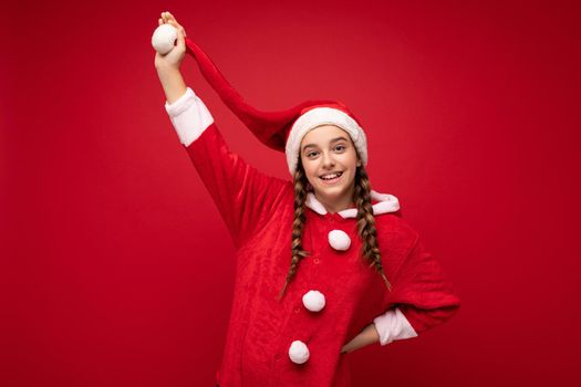 Photo shot of beautiful happy positive smiling brunette girl with pigtails wearing santa clause clothes isolated over red background wall looking at camera and having fun.