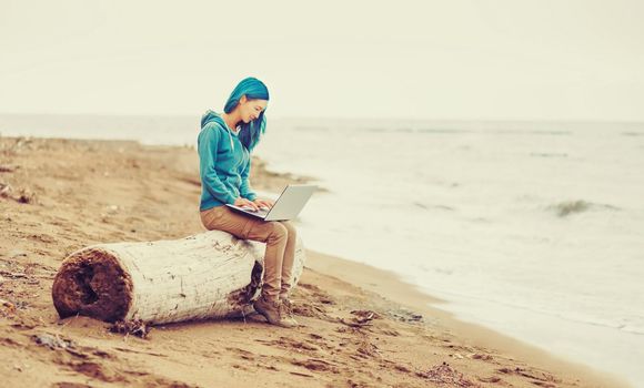 Freelancer young woman sitting on tree trunk and working on laptop on beach near the sea. Freelance concept