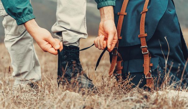 Hiker man tying shoelaces on nature outdoor, near backpack. View of legs. Hiking and leisure theme