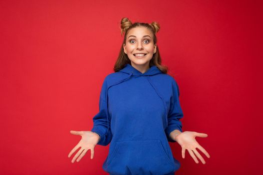 Young happy positive delightful attractive blond lady with two horns with sincere emotions wearing stylish bright blue hoodie isolated on red background with copy space.