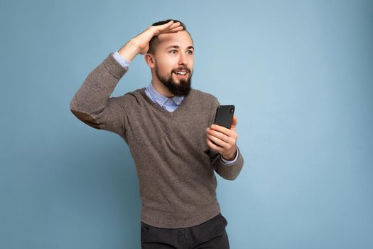 Handsome smiling brunette bearded man wearing grey sweater and blue shirt isolated on background wall holding mobile phone looking to the side.