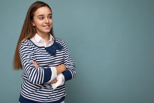 Shot of beautiful positive smiling brunette little girl wearing striped longsleeve standing isolated over blue background wall crossing hands and looking to the side. Copy space