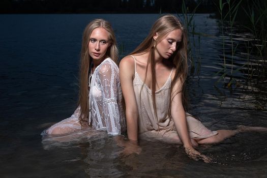 two attractive young twin sisters with long blond hair posing in light dresses in water of lake at summer night. stylish fashion photoshoot with flashlight. pretty models outdoors evening photosession