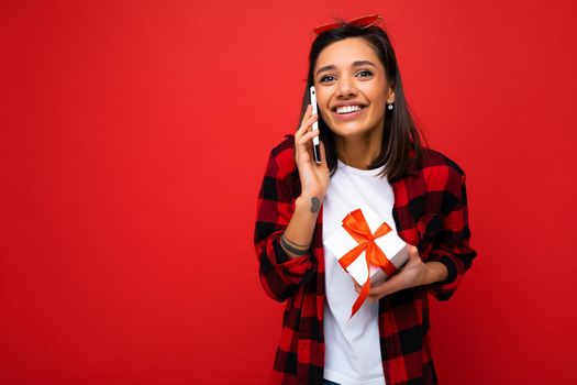 Shot of attractive positive smiling young brunette woman isolated over colourful background wall wearing everyday trendy outfit holding gift box and looking at camera. Copy space, mockup