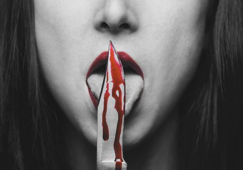Dangerous woman with red lips licking knife with red blood, monochrome image. Halloween or horror theme