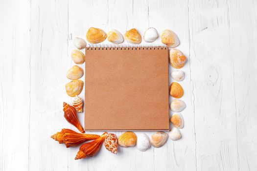 Open notepad with sea shells on white wooden background, top view
