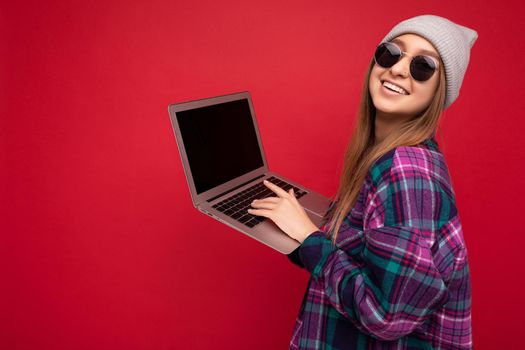 Side profile photo of beautiful smiling happy young woman holding computer wearing casual smart clothes sunglasses and hat looking at camera isolated on red background typing on keyboard text. Copy space