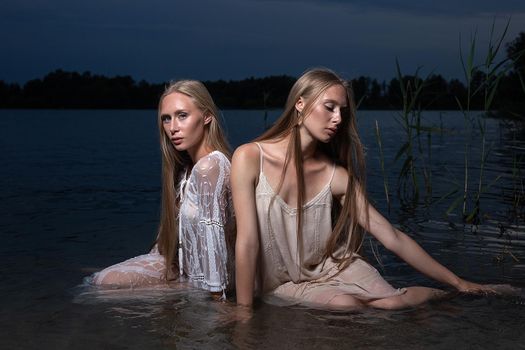 two young twin sisters with long blond hair posing in light dresses in water of lake at summer night, looking at camera. stylish fashion photoshoot with flashlight. outdoors evening photosession