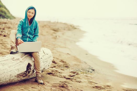 Freelancer girl sitting with laptop on tree trunk on beach near the sea and looking at camera. Freelance concept