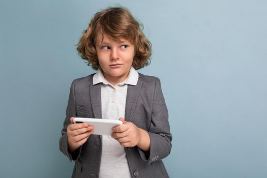 Photo shot of Handsome suspacted child boy with curly hair wearing grey suit holding and using phone isolated over blue background looking to the side playing games. Empty space