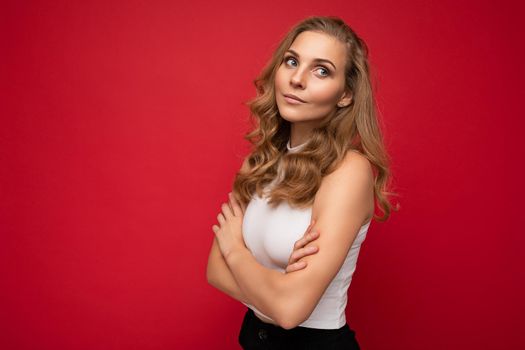 Portrait of young beautiful pretty self-confident thoughtful blonde businesswoman with sincere emotions wearing casual white top isolated over red background with empty space with arms crossed.