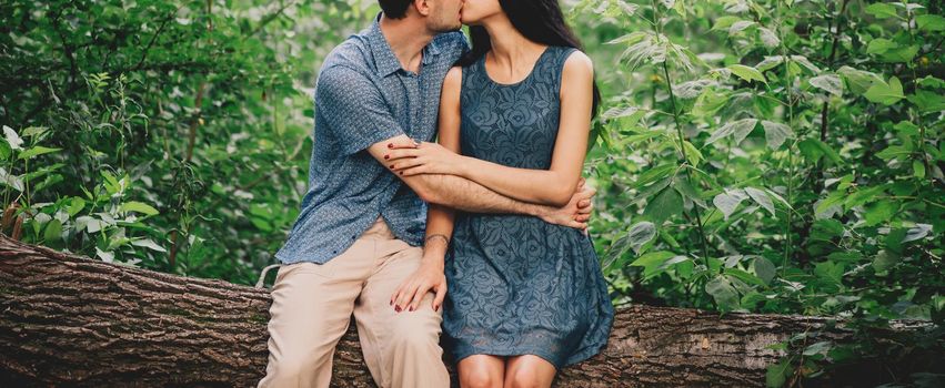 Young loving couple sitting on fallen tree trunk. Beautiful young woman kissing a handsome young man