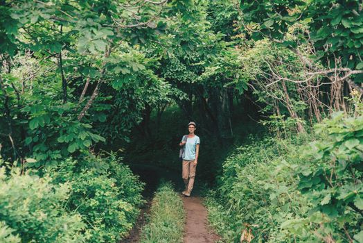 Traveler young woman walking in beautiful summer forest. Hiker girl goes out from the tunnel of green trees, girl looking at camera