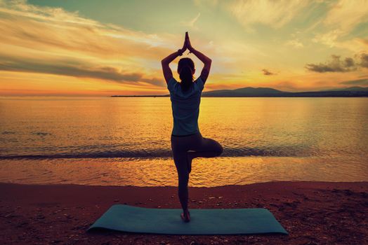 Woman doing yoga exercise in pose of tree on beach near the sea at sunset in summer, rear view