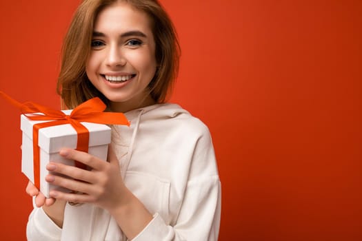 Beautiful happy young blonde woman isolated over colourful background wall wearing stylish casual clothes holding gift box and looking at camera. Empty space
