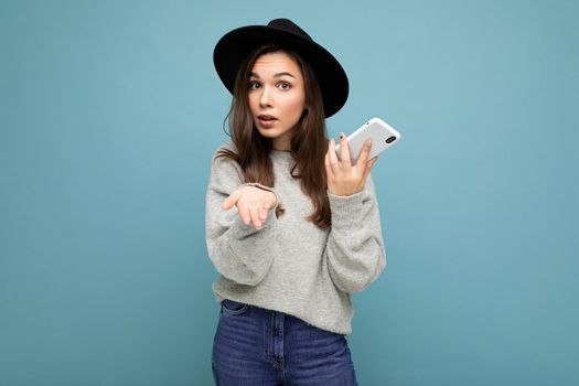 Beautiful young asking dissatisfied brunette woman pointon at camera wearing black hat and grey sweater holding smartphone looking at camera isolated on background.