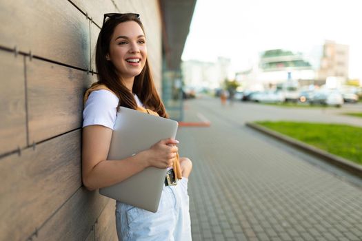 happy nice-looking charming attractive lovely fascinating cheerful cheery straight-haired brunet woman looking to side in the street with laptop computer and glasses in hands with white t-shirt and jeans.