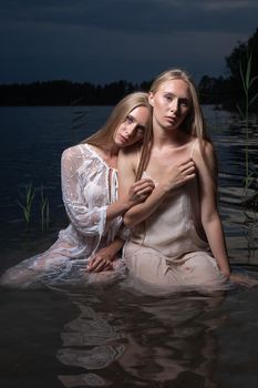 two young twin sisters with long blond hair posing in light dresses in water of lake at summer night, looking at camera. stylish fashion photoshoot with flashlight. outdoors evening photosession