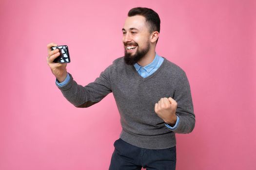 Shot of happy handsome young brunette unshaven man with beard wearing everyday grey sweater and blue shirt isolated on background wall holding smartphone watching videos via mobile phone looking at phone screen display and celebrating winning.