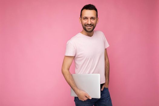 Handsome smiling brunet man holding laptop computer looking at camera in t-shirt on isolated pink background.