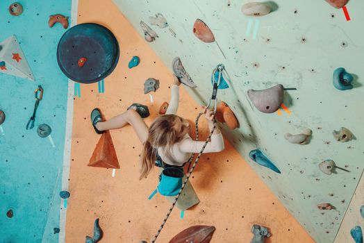 Girl wearing in safety equipment climbing on artificial boulders in gym. Girl inserting the rope in a quickdraw