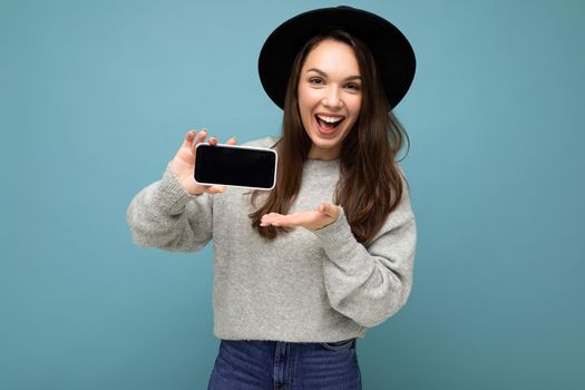 Pretty young smiling woman wearing black hat and grey sweater holding phone looking at camera isolated on background.Mock up, cutout