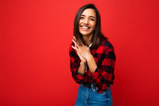 Photo of young beautiful happy smiling brunette woman wearing trendy white t-shirt and red check shirt . Sexy carefree female person posing isolated near red wall in studio with free space. Positive model with natural makeup.