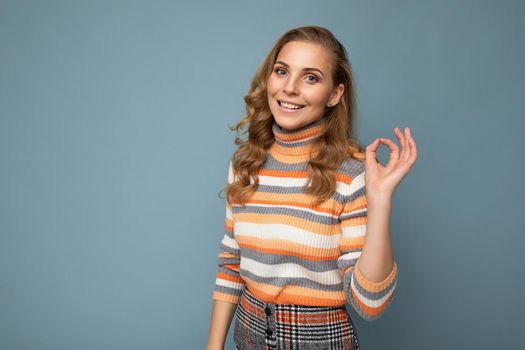Portrait of young winsome attractive happy smiling blonde woman with wavy-hair wearing striped sweater isolated over blue background with empty space and showing ok gesture.